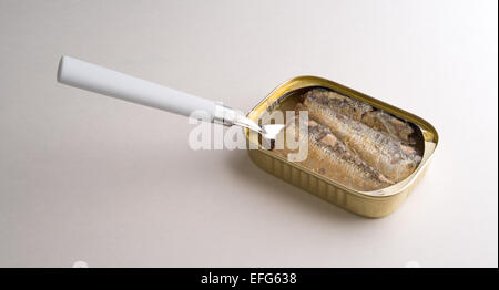 An opened can of sardines in oil and water with a fork inserted in the tin on a white table top illuminated by window light. Stock Photo