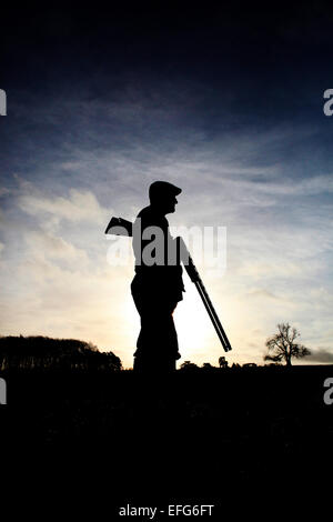Silhouetted male game shooter standing with shotgun against sunset sky