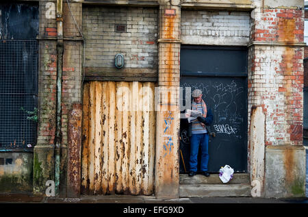 Man in a flat cap reads the newspaper in the doorway of a dilapidated street of Manchester city centre. Editorial use only. Stock Photo