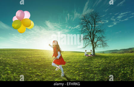 Happy girl running in a meadow with colorful balloons on a background of parents Stock Photo