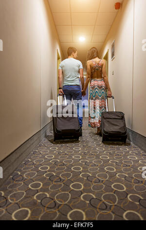 Young couple standing at hotel corridor upon arrival, looking for room, holding suitcases. Stock Photo