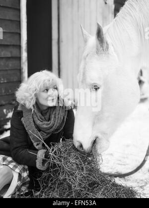 Attractive blond woman feeds a white horse, overcast winter day, black and white image, focus on horse eyes Stock Photo