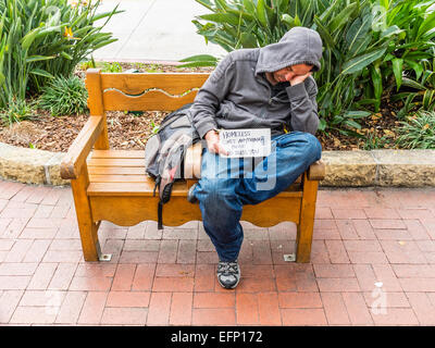 A homeless man sleeps outside on a sidewalk bench on State Street in Santa Barbara, California while holding a begging sign. Stock Photo