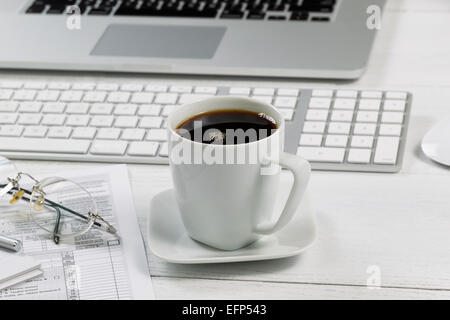 Close up of a fresh cup of black coffee with laptop, keyboard, pen, mouse, reading glasses and tax forms in background on white Stock Photo