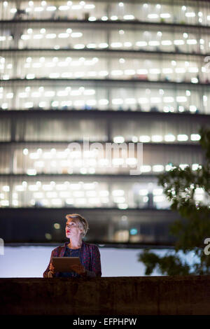 Mature businesswoman using digital tablet in front of office building at night, London, UK Stock Photo