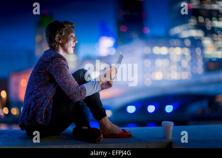 Mature businesswoman sitting on waterfront using  digital tablet at night, London, UK Stock Photo