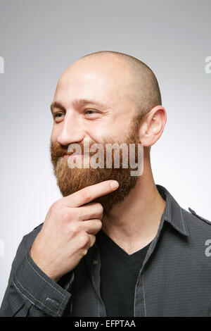 Studio portrait of mid adult man with shaved hair stroking overgrown beard Stock Photo