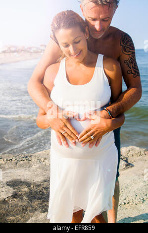 Pregnant mature woman and husband making heart shape on stomach at beach Stock Photo