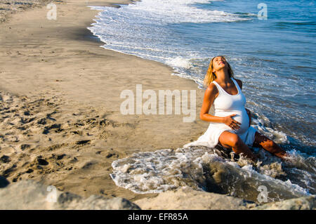 Pregnant mature woman sitting in ocean waves on beach whilst touching stomach Stock Photo