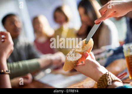 A group of people sitting at a table, eating and chatting. A woman buttering a slice of baguette. Stock Photo