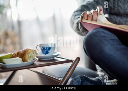 Man balancing a book on his knee cup and saucer and a plate apple and croissant next to him. Stock Photo