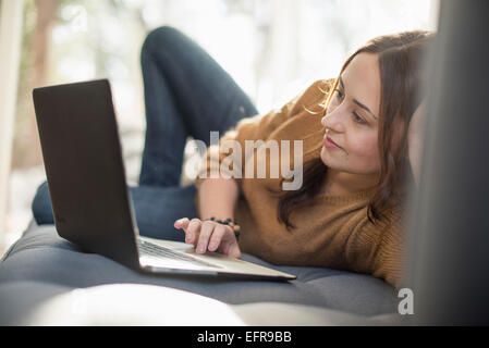 Woman lying on a sofa looking at her laptop, smiling. Stock Photo