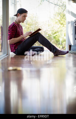 Man wearing a baseball cap backwards, sitting on the floor in a living room, looking at a digital tablet. Stock Photo