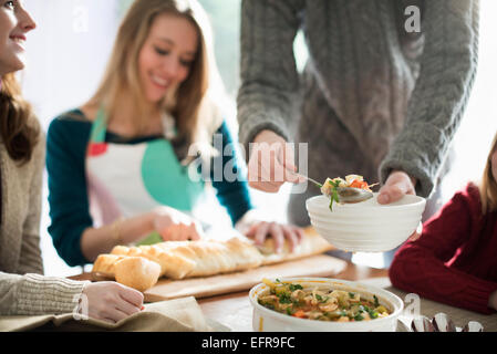 People sitting and standing at a table, a man serving food into a bowl, a woman slicing a baguette. Stock Photo