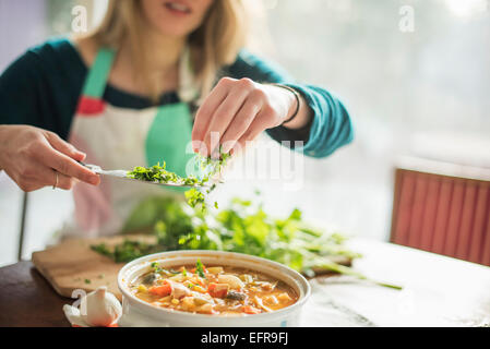 A woman wearing an apron, sitting at a table, sprinkling herbs into a bowl of vegetable stew. Stock Photo
