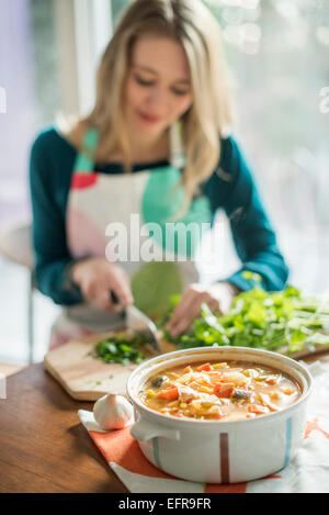 A woman wearing an apron, sitting at a table, chopping herbs, a bowl of vegetable stew in the foreground. Stock Photo