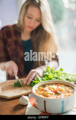 A woman sitting at a table, chopping herbs, a bowl of vegetable stew in the foreground. Stock Photo