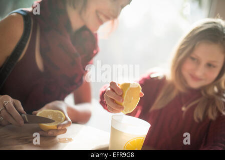 A woman and a girl sitting at a table, girl squeezing the juice from a lemon into a glass. Stock Photo