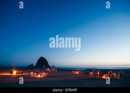Crowds of people sitting by glowing campfires on Cannon Beach at dusk. Haystack Rock in the background. Stock Photo