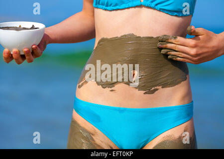 Young slim woman smearing mud mask on the body on the beach Stock Photo