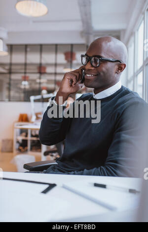 Happy young african man sitting at his desk talking on his mobile phone in office. African executive using cell phone. Stock Photo
