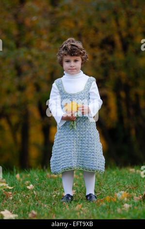 Young female child playing with autumn leaves on green lawn outside wearing a cute crocheted dress Stock Photo