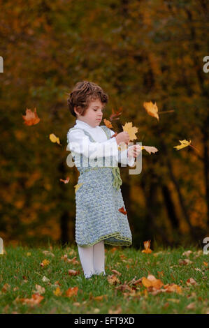 Young female child playing with autumn leaves on green lawn outside wearing a cute crocheted dress Stock Photo