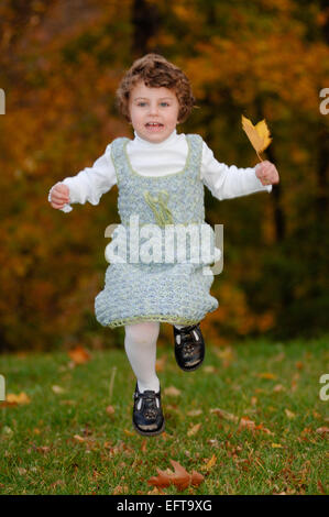 Young female child playing with autumn leaves on green lawn outside wearing a cute crocheted dress Stock Photo