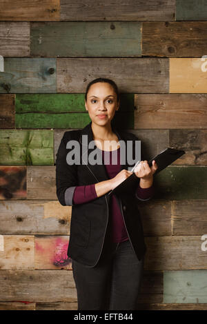 Portrait of confident young african businesswoman standing against wooden wall in office holding a clipboard. Stock Photo