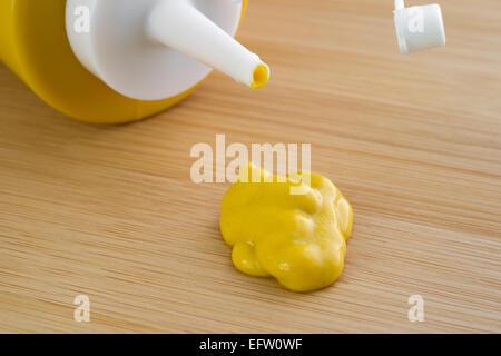 Close view of a bottle of mustard laying on its side with a small blob squeezed out in front onto a wood cutting board. Stock Photo