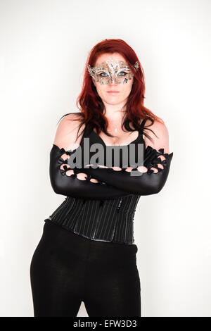 A young redhead red haired woman goth girl wearing a black corset and  long black elbow-gloves and a silver mask with her arms crossed folded Stock Photo