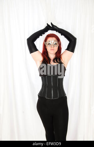 A young redhead red haired woman goth girl wearing a black corset and  long black elbow-gloves and a silver mask with her arms raised above her head Stock Photo