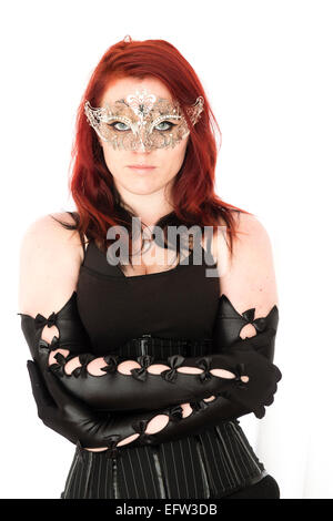 A young redhead red haired woman goth girl wearing a black corset and  long black elbow-gloves and a silver mask with her arms crossed folded facing ahead looking direct Stock Photo