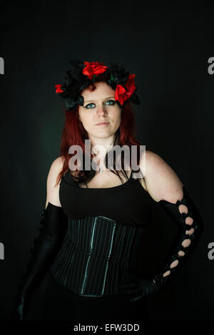 A young redhead red haired woman goth girl wearing a black corset and  long black elbow-gloves and red flowers in her hair Stock Photo