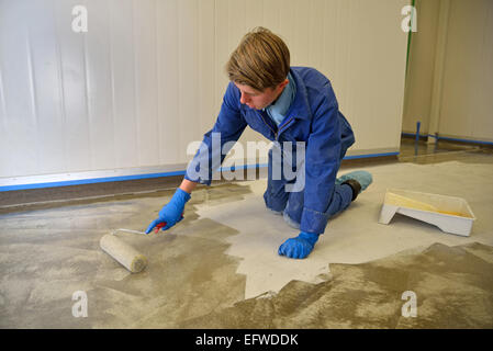 An apprentice tradesman uses a roller to apply an epoxy product to the floor of an industrial building Stock Photo