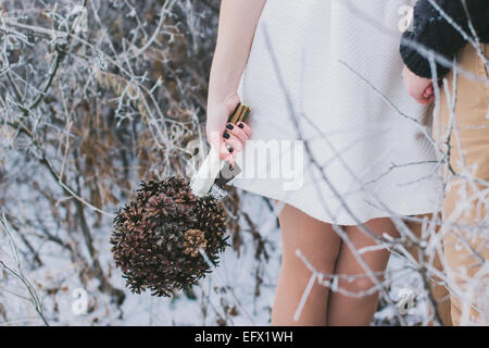Bride and groom holding hands in snow-covered winter forest; bride holding wedding bouquet made of pine cones Stock Photo