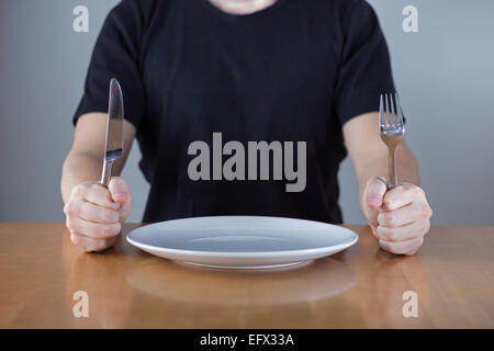 An unrecognizable man wearing black shirt sitting at a table in front of an empty plate waiting for food, holding fork and knife Stock Photo