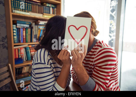 Flirty couple sitting in library behind paper with red heart Stock Photo