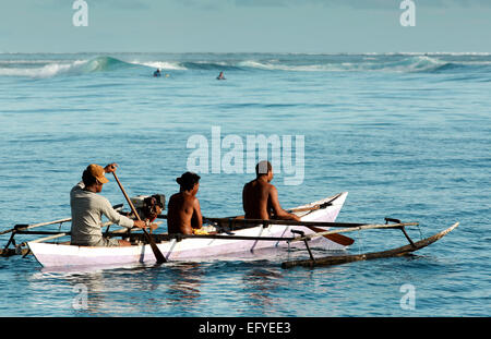 Fishermen in wooden outrigger canoe paddle past tourists surfing in the waves in the Maluku Islands, Indonesia Stock Photo