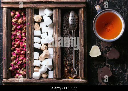 Set of dry black tea, tea roses and sugar cubes in wooden box with vintage tea strainer and cup of tea Stock Photo