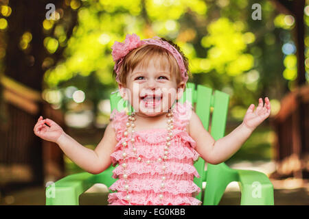 Happy girl in a party dress sitting in garden chair laughing Stock Photo