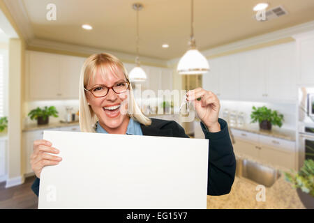 Happy Young Woman Holding Blank Sign and Keys Inside Beautiful Custom Kitchen. Stock Photo