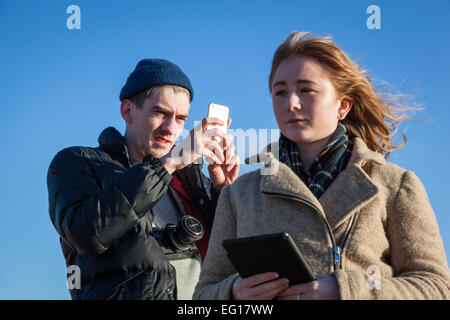 Young man wearing 'puffa' jacket and woollen hat in winter uses his iphone take a photograph of a girl with red hair . Stock Photo