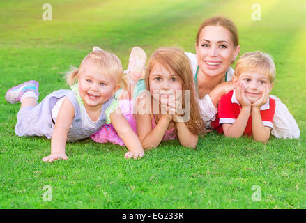 Happy big family having fun outdoors, young mother with three cute kids lying down on fresh green field, love and togetherness Stock Photo