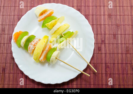 Delicious fruit skewers on a white plate placed on a bamboo tablecloth Stock Photo