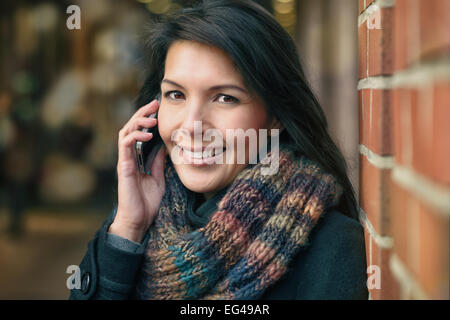 Smiling Young Woman in Autumn Fashion Talking on Phone While leaning against a red brick pillar Stock Photo