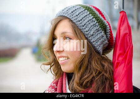 Young brunette wearing a hand made woolen hat and a red umbrella outdoors Stock Photo