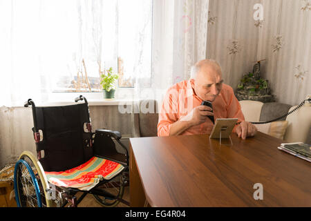 Sitting Elderly Man Shaving Beards with Electric Razor at the Living Room with his Wheel Chair on the Side. Stock Photo