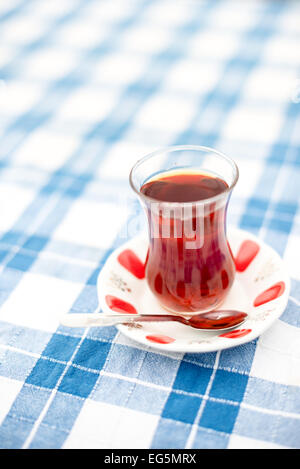 A glass of Turkish tea on a checkered blue and white tablecloth in Istanbul, Turkey. Turkish tea is traditionally served in small glasses shaped like this on a small plate. It is a black tea taken without milk but sometimes with beet sugar. Stock Photo