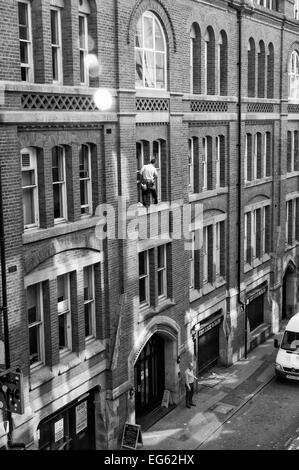 Window cleaner on very narrow ledge 3storeys up with no safety gear and man below oblivious to what is above him Stock Photo